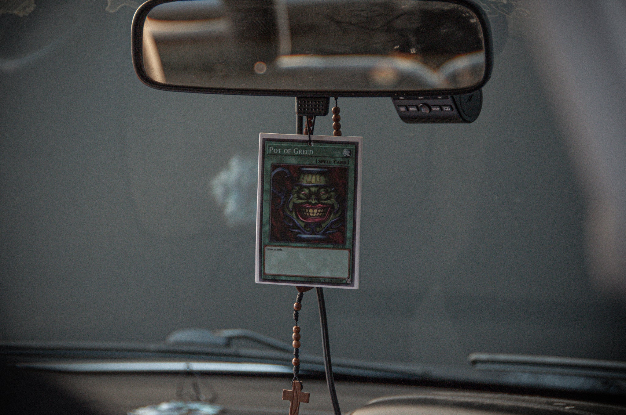 Hanging from the rearview mirror of a car, an "Incompletegl Pot of Greed! Air Freshener," featuring a grinning, green-faced character, is displayed alongside a rosary with a crucifix. In the background, part of the car dashboard and a blurred view through the windshield can be seen.