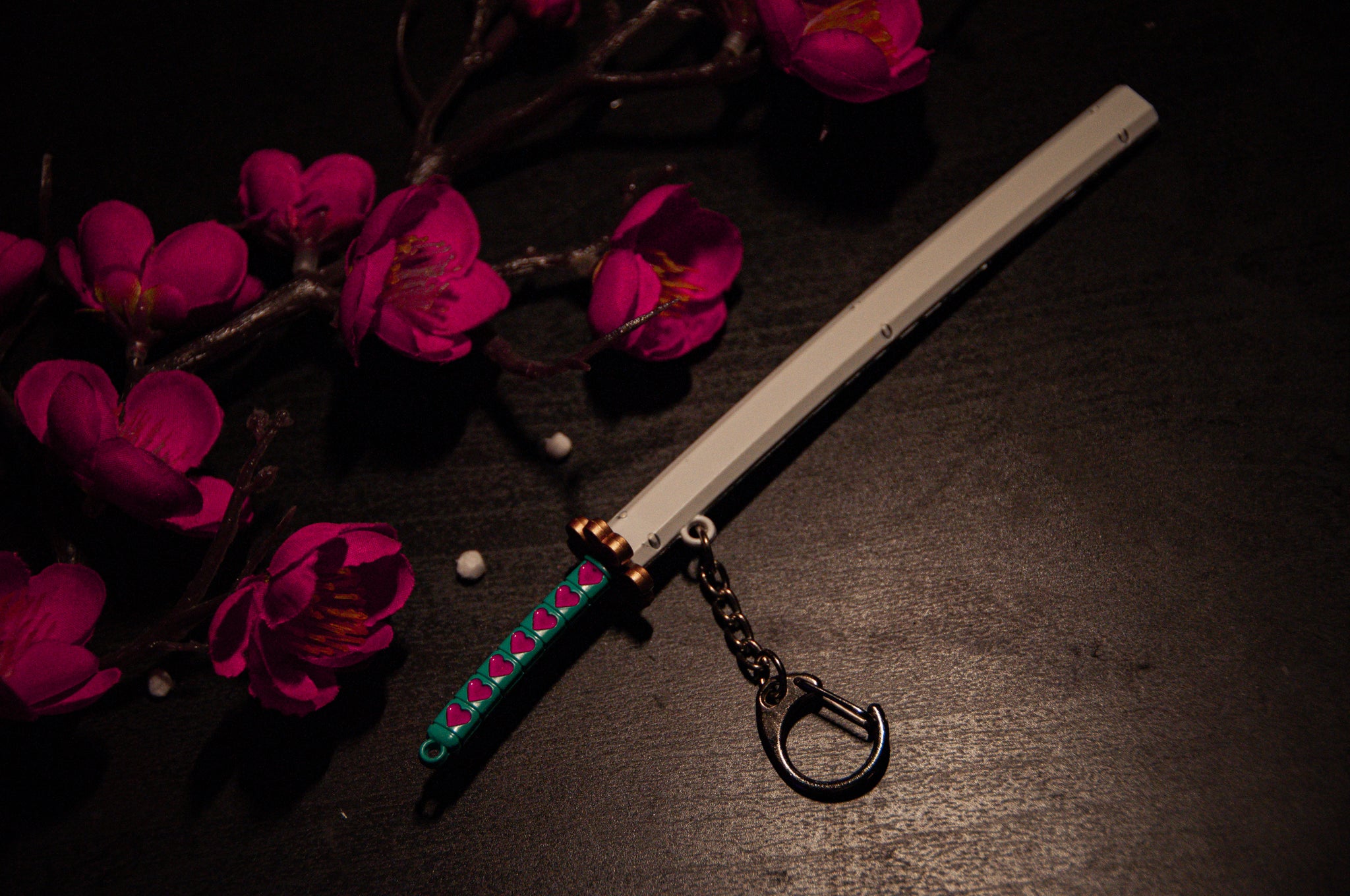 The Incompletegl Demon Slayer Swords! Keychain, featuring a decorative design with a green and pink handle, white gravity-sheath, and gold guard, is placed beside a branch of vibrant pink cherry blossoms on a dark surface, making it perfect for anime enthusiasts.