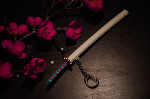 The Incompletegl Demon Slayer Swords! Keychain, featuring a decorative design with a green and pink handle, white gravity-sheath, and gold guard, is placed beside a branch of vibrant pink cherry blossoms on a dark surface, making it perfect for anime enthusiasts.