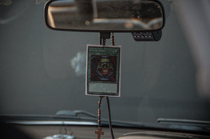 An interior car rearview mirror adorned with an iconic "Pot of Greed" Yu-Gi-Oh! Spell Card and a rosary ending in a cross, along with a watermelon-scented air freshener from the Ultimate Deck! | Air Freshener Bundle by Incompletegl. The background showcases a blurred view of the car's dashboard and windshield.