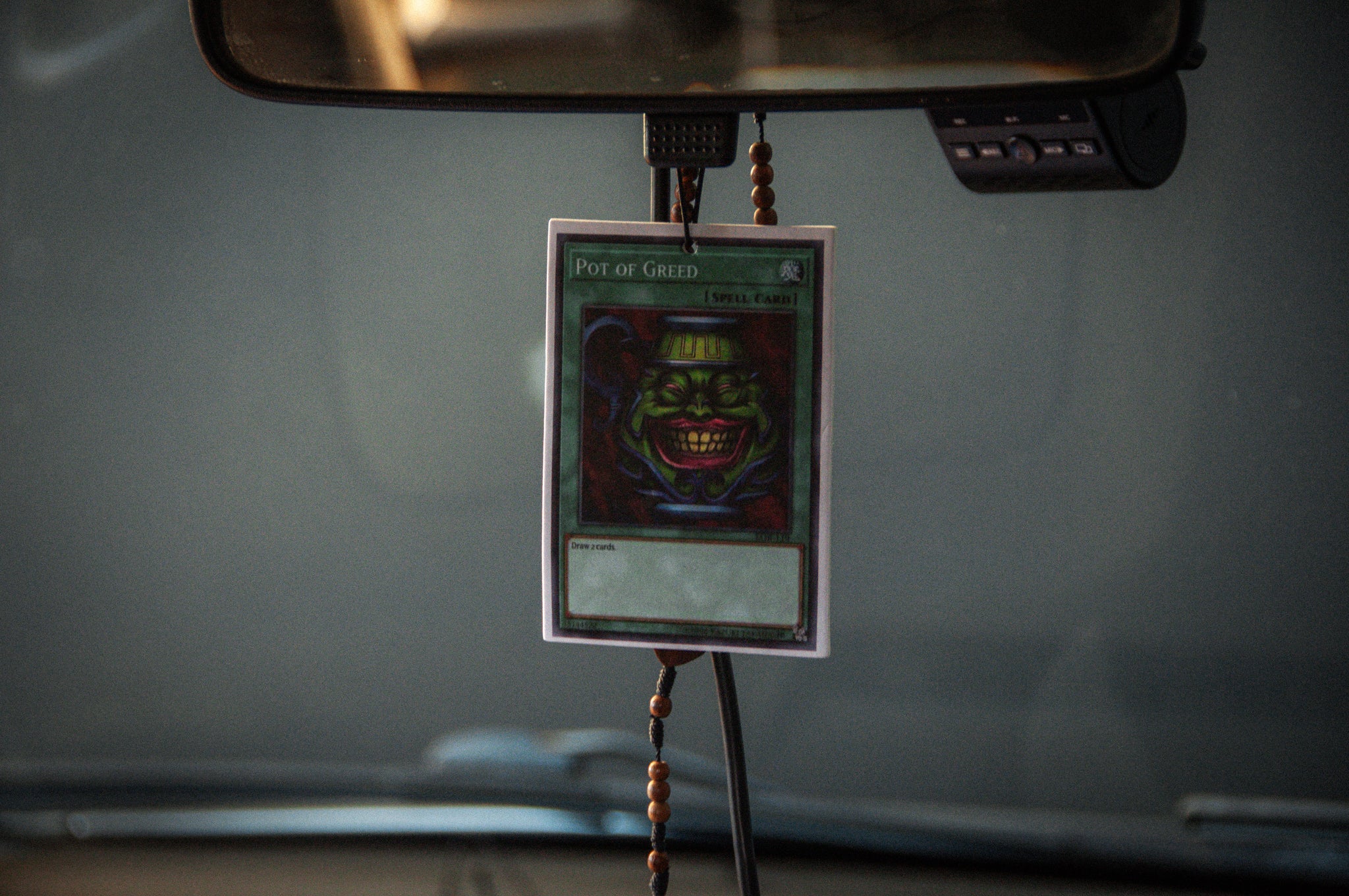 An "Incompletegl Pot of Greed! Air Freshener" is hanging from the rearview mirror of a car. The air freshener features a grinning green pot with beady eyes. The car's dashboard and a blurred background are partially visible.