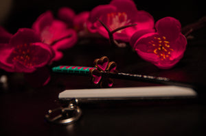 A close-up photograph showcases vibrant pink cherry blossoms in the background. In the foreground, there is a colorful pencil with a heart-shaped eraser, a closed notebook, and a keychain styled after Demon Slayer Swords! from the brand Incompletegl. The scene is warmly lit with a dark backdrop.