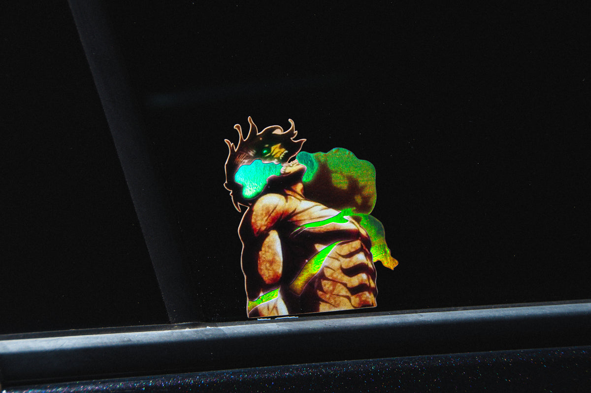 A sticker on a car window, called the "Eren Yeager, Titan Form! | Holo Peeker Decal" by Incompletegl, depicts a muscular figure with spiky hair and glowing green eyes, reminiscent of Eren Yeager in his Titan form, holding a reptilian creature in a dramatic pose. The dark background emphasizes the vibrant colors of the characters.
