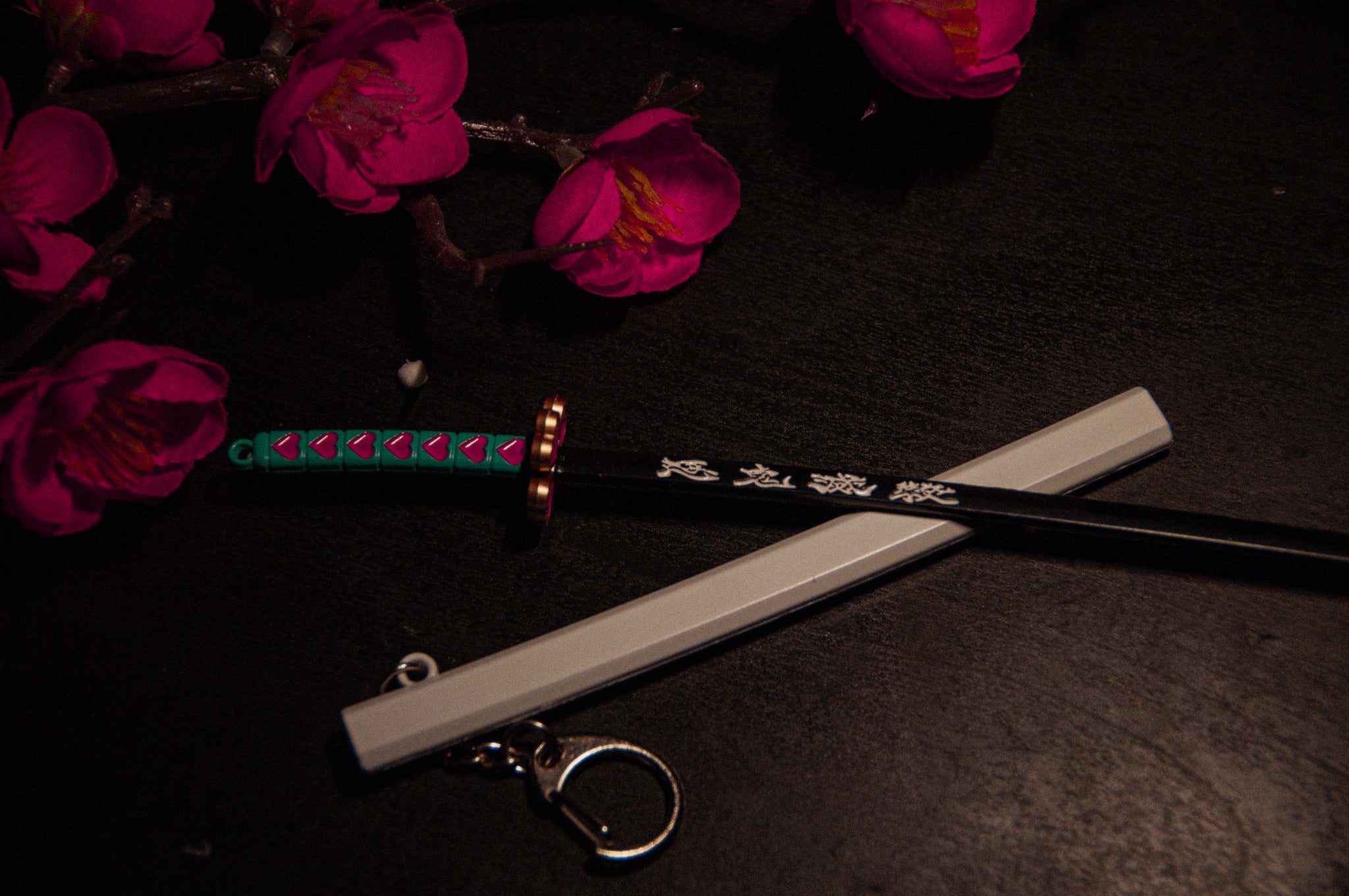 On a dark surface lies a detailed miniature katana from the Demon Slayer Swords! | Keychains collection by Incompletegl, featuring a vibrant handle and accompanied by a white sheath. Surrounding the katana are bright, artificial cherry blossom flowers that add a decorative touch to the scene—a perfect piece for anime enthusiasts and collectors of Demon Slayer Sword Keychains.