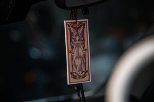 A rectangular Cardcaptor Sakura air freshener by Incompletegl, titled 'The Nothing,' features an illustration reminiscent of a Clow Card, depicting a mystical, robed female figure with antler-like horns. Hanging by a beaded string from a rearview mirror inside a car, the words "THE ANTLER" are visible at the bottom.