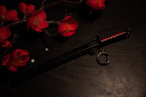 An exquisitely crafted keychain lies on a dark surface surrounded by red cherry blossom flowers, featuring a sheathed black sword with a red and black handle and adorned with a crescent moon charm—an ideal addition for anime enthusiasts who appreciate elegant details. Introducing the "Demon Slayer Swords! | Keychains" by Incompletegl.