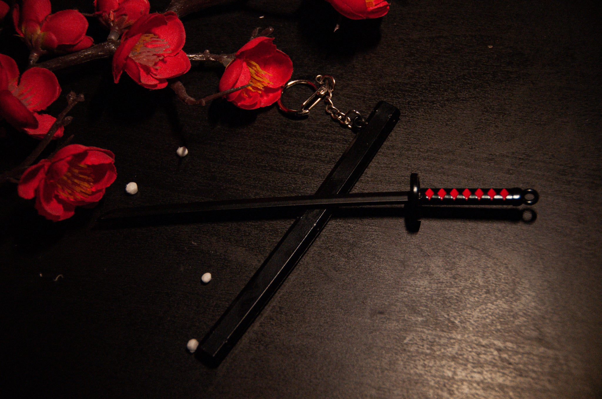 An Incompletegl Demon Slayer Swords! keychain, featuring a mini black katana with a red and black hilt, rests on a dark surface. Nearby, small white beads and a branch adorned with red flowers provide a striking contrast. The dimly lit background accentuates the keychain, making it an ideal accessory for anime enthusiasts and fans of Demon Slayer Sword Keychains.