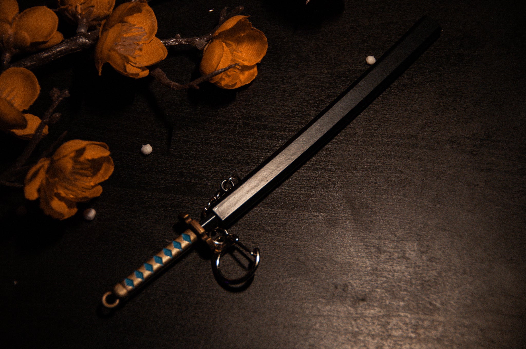 A sleek, rectangular sword keychain from the Demon Slayer Swords! collection by Incompletegl, featuring a blue and white checkered handle, lies on a dark surface. The background is adorned with vibrant orange artificial flowers and two small white beads scattered nearby. A perfect accessory for anime enthusiasts.