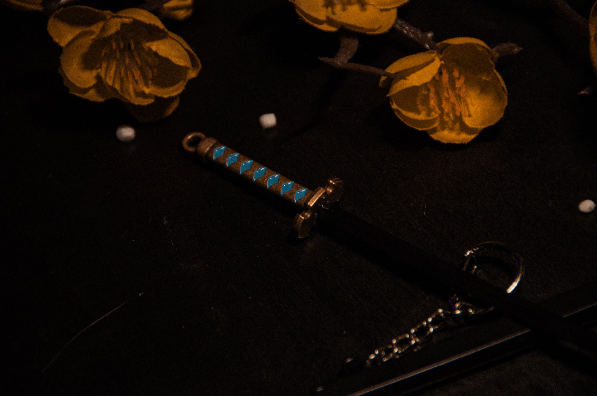 An intimate photograph captures a keychain from the "Demon Slayer Swords!" collection by Incompletegl, featuring a miniature sword adorned with a blue and gold hilt resting on a dark surface. Beside the keychain are intricately detailed orange flowers. The dim lighting accentuates the vibrant colors and textures of both the sword and the flowers, delighting anime enthusiasts.