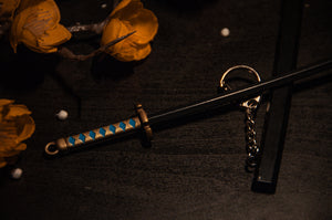 A close-up of an Incompletegl Demon Slayer Swords! keychain reveals a miniature samurai sword with a detailed handle showcasing blue and white diamond patterns. The sword, reminiscent of popular Demon Slayer Sword Keychains, is laid on a dark surface beside vibrant orange flowers. The sheath is also visible, resting parallel to the blade.