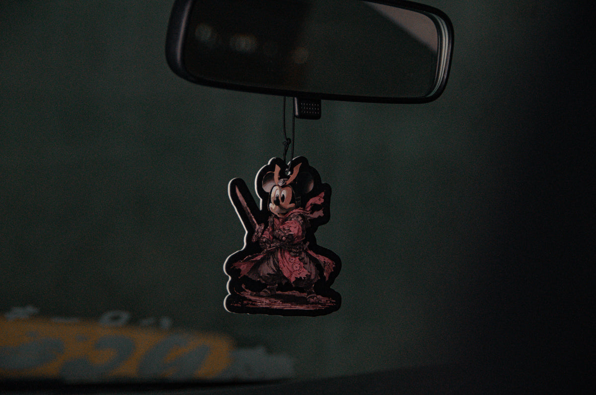 A dark, moody photo of a car's rearview mirror adorned with an air freshener that showcases a stylized, heavily-detailed character inspired by the DONALD DUCK x RONIN SUIT Oversized T-Shirt from Incompletegl. The character is dressed in traditional samurai armor and wields a katana.