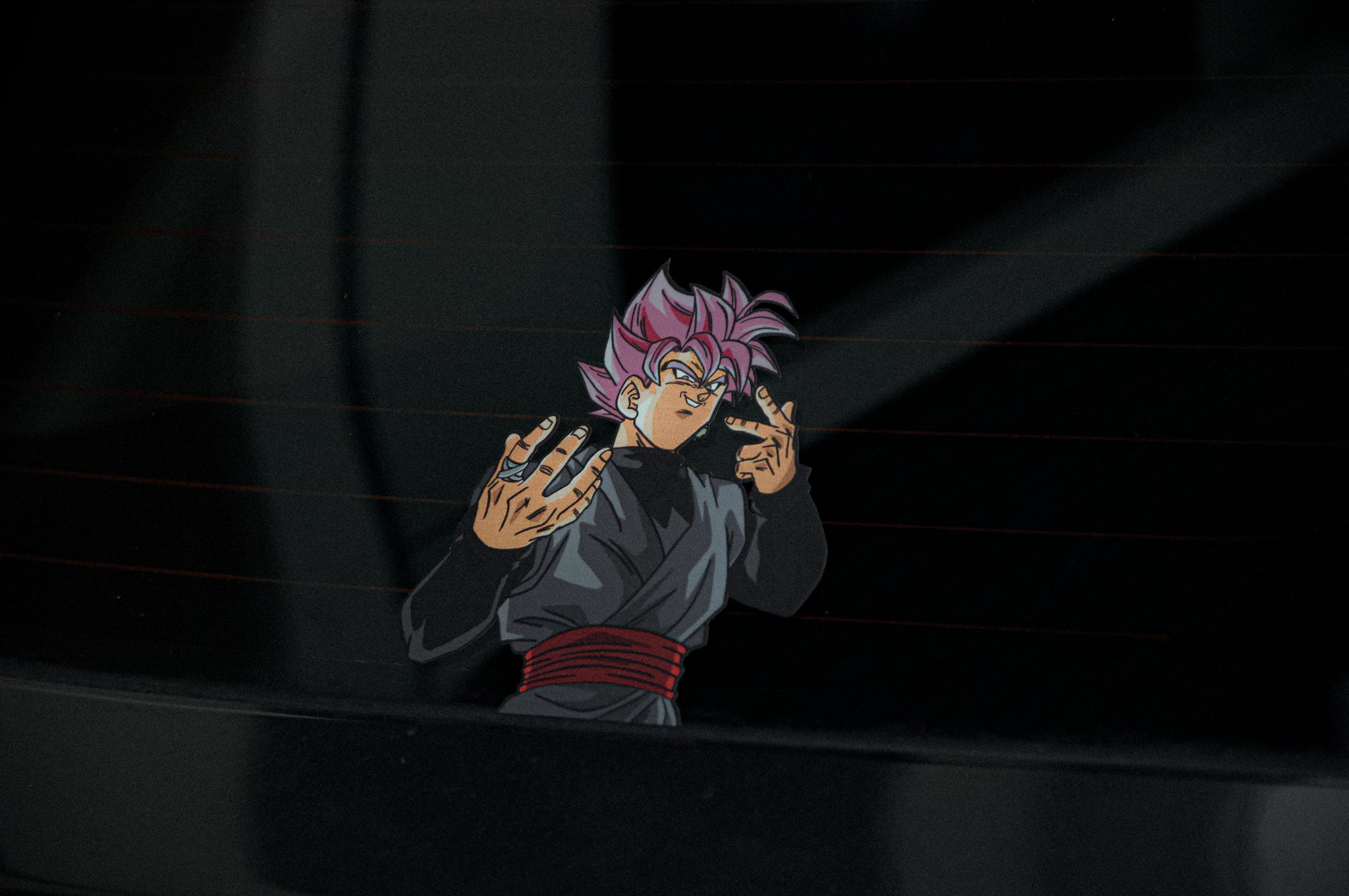 A vibrant Dragon Ball Z Super Pack! sticker from Incompletegl featuring an anime character with pink spiky hair, evoking Saiyan spirit and wearing a black outfit and red belt, is placed on a car window. The character is making a hand gesture with both hands and has a serious expression. The background of the image is dark and reflective.