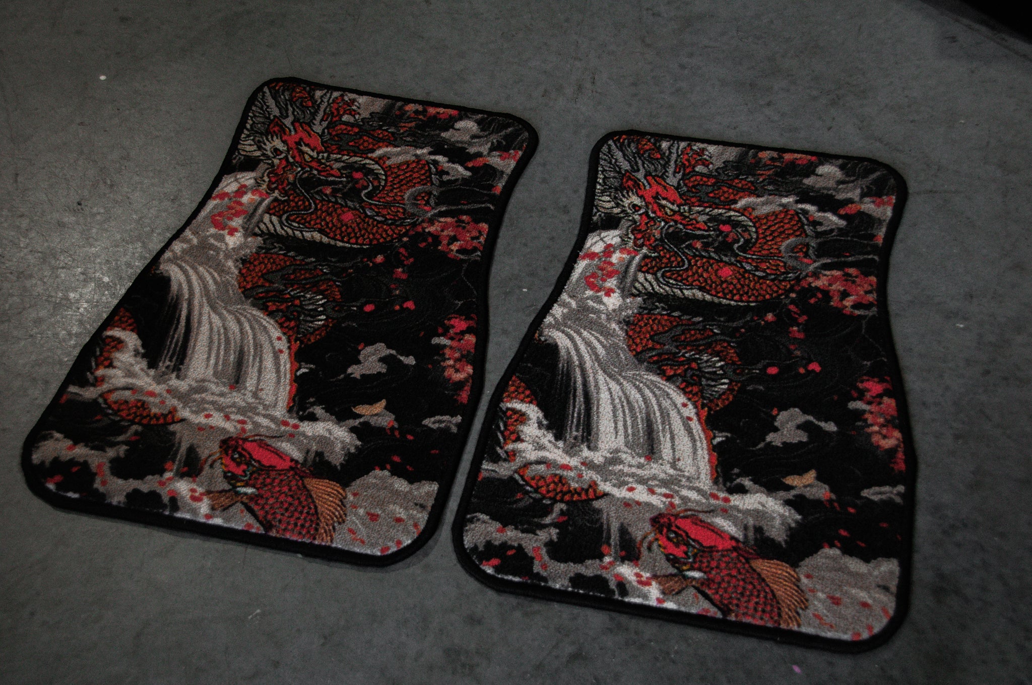 The Dragon x Koi floor mats by Incompletegl showcase an intricate design featuring two black JDM mats adorned with red koi fish swimming around Dragon’s Gate, set against a gray backdrop of cascading waterfalls and cloudy floral patterns.