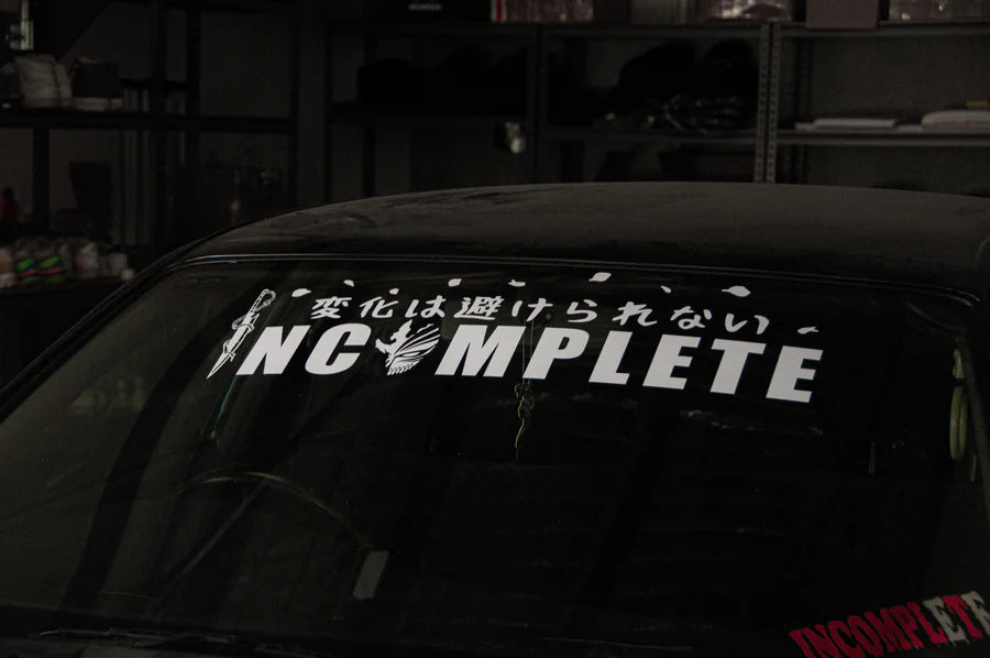 In a dimly lit garage, an Incompletegl car windshield showcases large white text of the brand name "INCOMPLETE," surrounded by Japanese characters. The setting resembles the Bleach Mega Mystery! Anime Bundle Pack, enticing curiosity like a scene from an anime-themed mystery collection waiting to unravel its secrets among shelves filled with various curiosities.