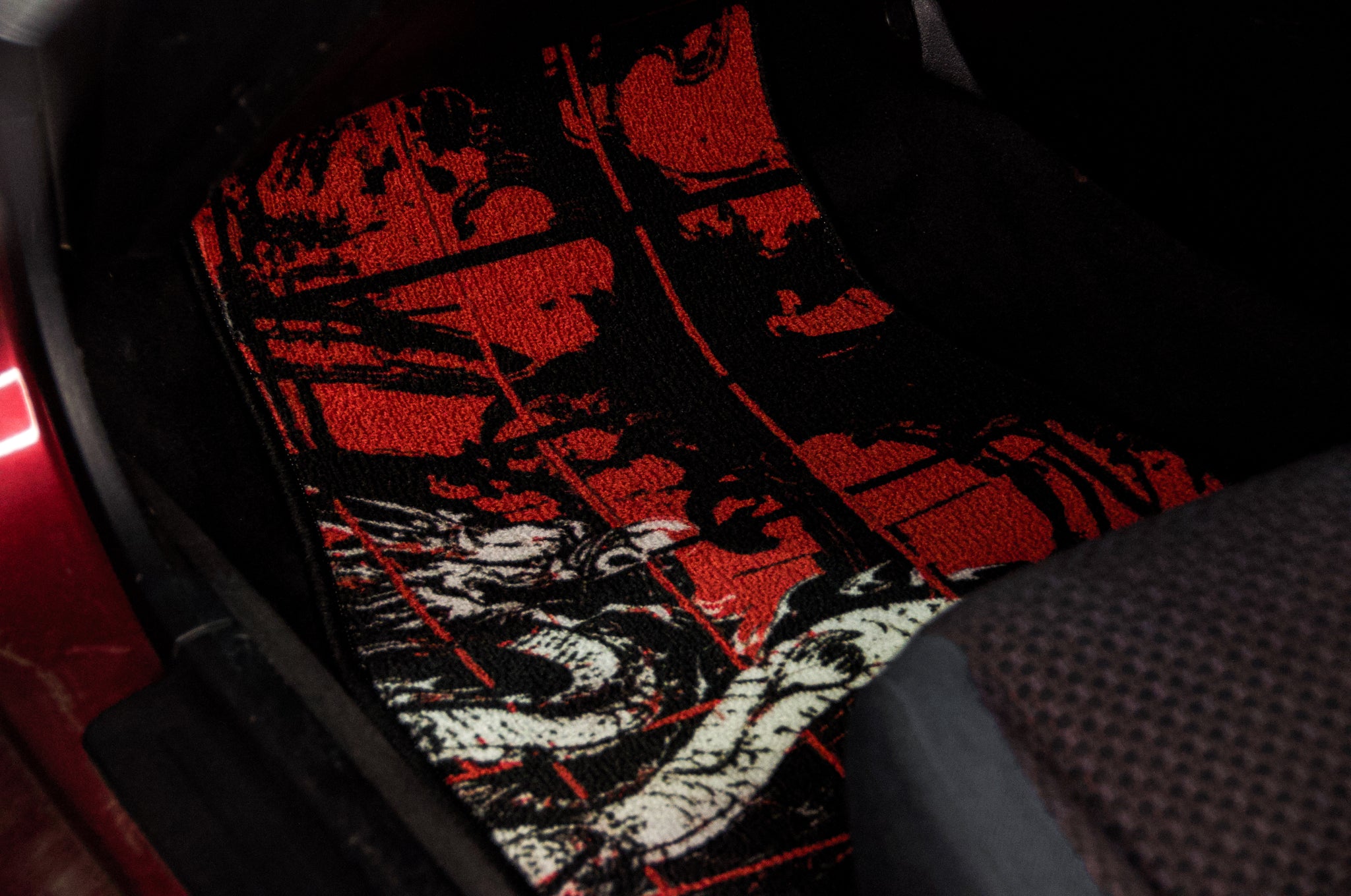 In the vehicle’s footwell area, the Chaos Red Checkered! | Floor Mats by Incompletegl, featuring a traditional Japanese dragon design, are displayed. The mat, with its black background and intricate red and white dragon depiction, seamlessly integrates traditional Japanese art into your ride. The surrounding interior of the car includes part of the seat and the car's floor.