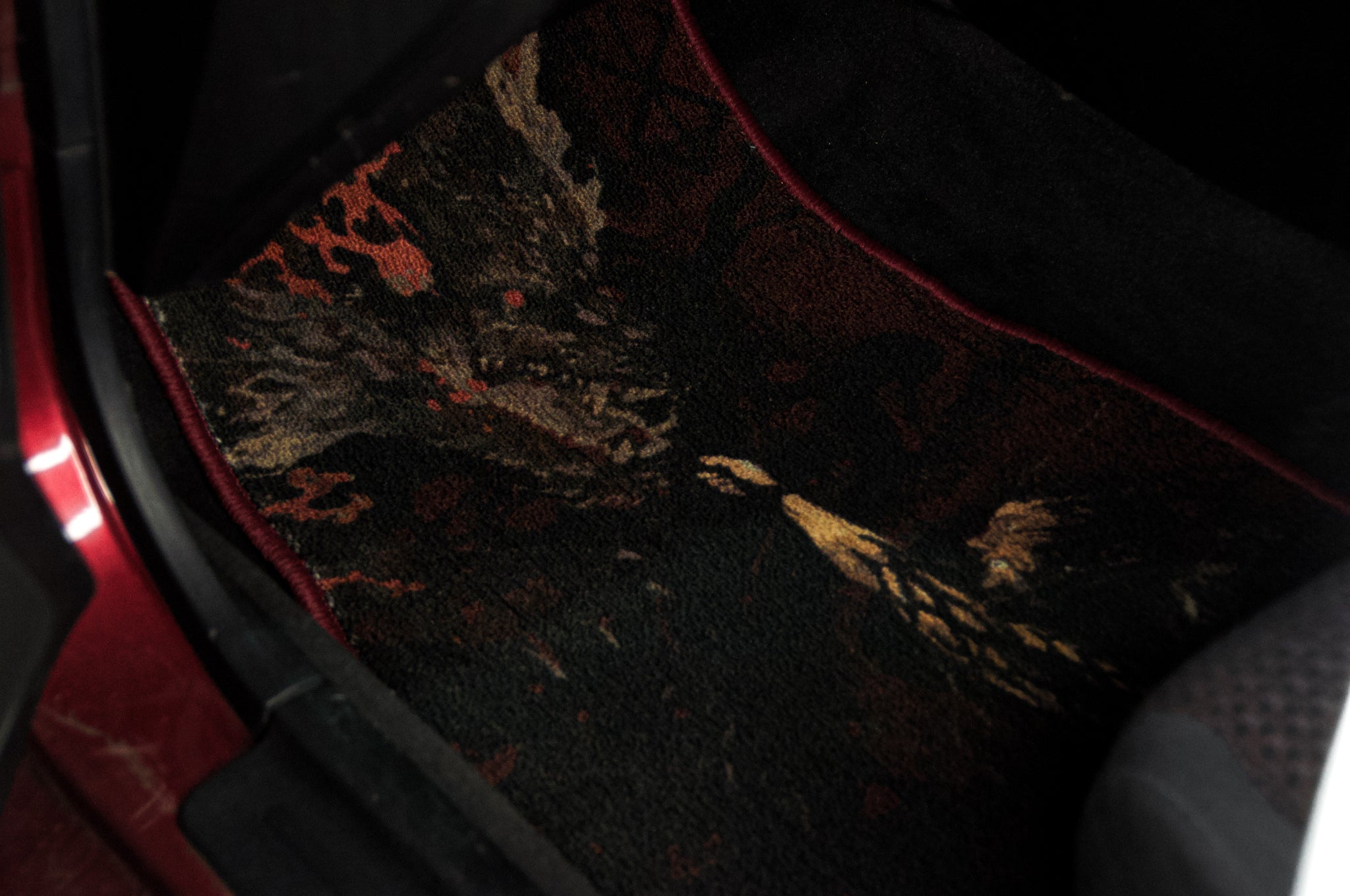 A close-up of an Incompletegl Berserk!!! floor mat showcasing a detailed dragon design with its head turned to the side, enveloped by dark, fiery elements. The dimly lit car interior accentuates the vibrant and intricate manga art-inspired design on the mat.