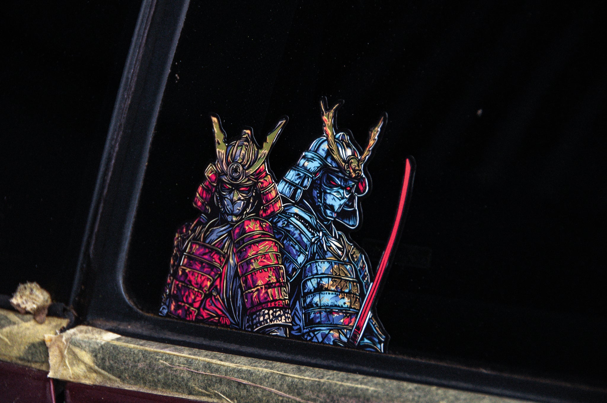 The car window is adorned with the "Samurais x Red & Blue! | Holographic Peeker Decal" by Incompletegl, showcasing a vibrant image of two samurai warriors in red and blue armor. Each warrior wields a sword and wears a horned helmet, capturing the spirit of Japanese culture, while the window frame exhibits slight wear.