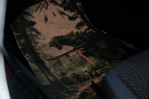 The Godzilla x Torii Shrine Floor Mats! by Incompletegl showcase a Japanese-themed design adorned with a Torii Shrine, trees, and a dragon-like creature evocative of Godzilla. The mat is positioned inside a vehicle, partially visible under the seat, with muted shades of green, brown, and red.