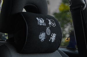 A car seat with Incompletegl's Gear 5!!! headrest pillow, in black, featuring white embroidered designs inspired by Luffy's evolution. The background shows a blurred view of greenery through the car window.