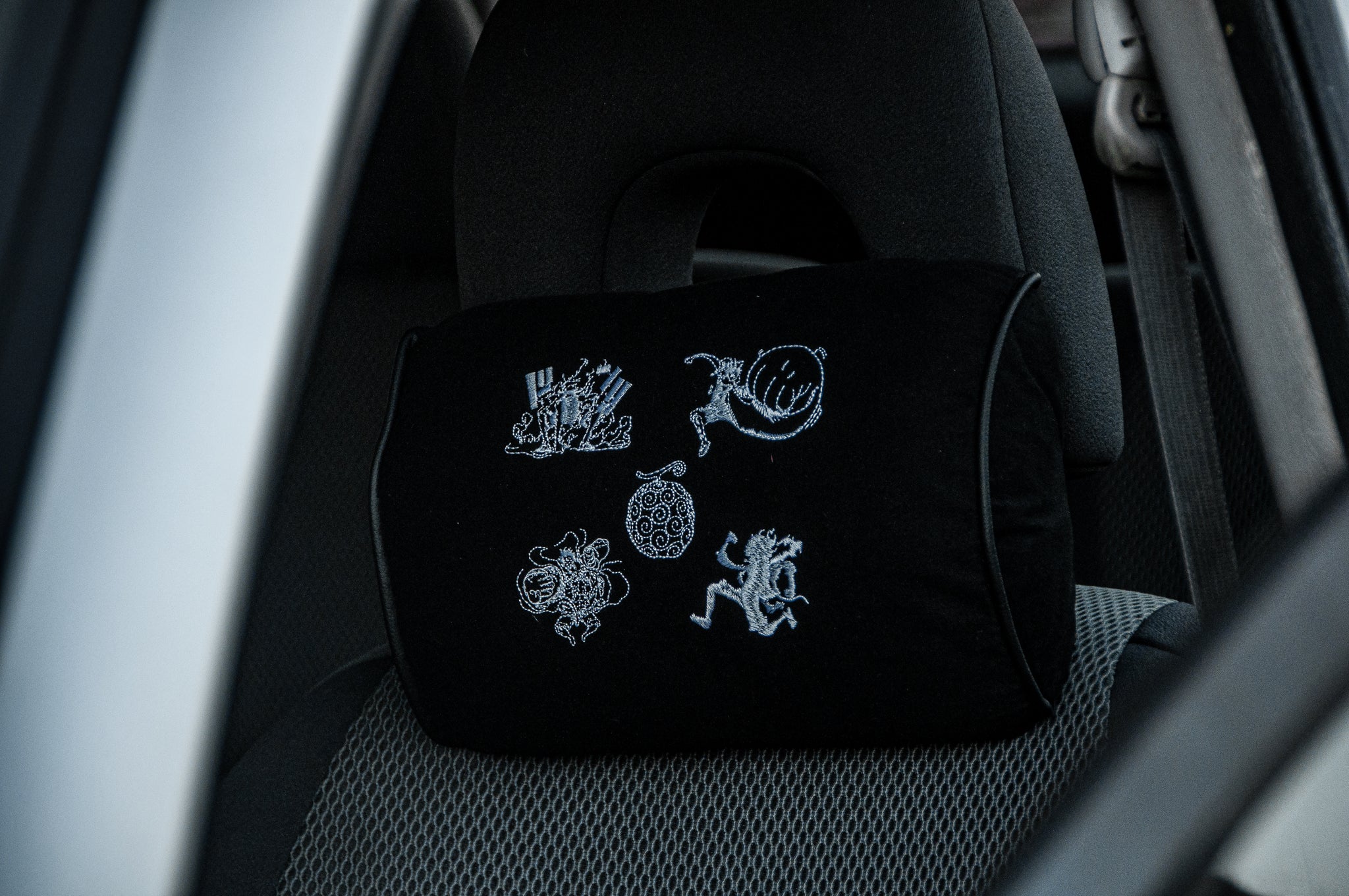 A black headrest pillow by Incompletegl, featuring white embroidered abstract designs and a figure, sits on a car seat. Named "Gear 5!!! | Headrest Pillow," it adds character to the grey interior visible through the open door.