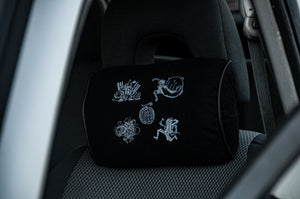 A black headrest pillow by Incompletegl, featuring white embroidered abstract designs and a figure, sits on a car seat. Named "Gear 5!!! | Headrest Pillow," it adds character to the grey interior visible through the open door.