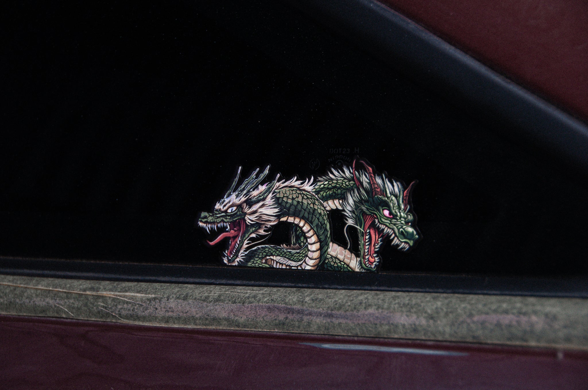 The rear window of a JDM car is adorned with the Twin Dragons | Holographic Peeker Decal from Incompletegl, showcasing a twin-headed Japanese dragon with intricate scales and fierce expressions. The decal displays vibrant green and white hues against the dark tinted glass background.