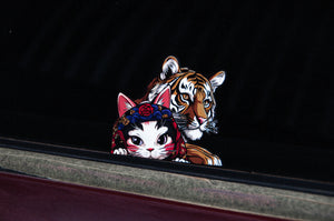 The "Neko & Lion! | Holographic Peeker Decal" by Incompletegl features a cartoon-style image of a neko wearing a floral headband, peeking mischievously over a ledge, with a realistic tiger lurking behind. Set against a black background, the slightly visible edge of the car window enhances the decal's intriguing effect.