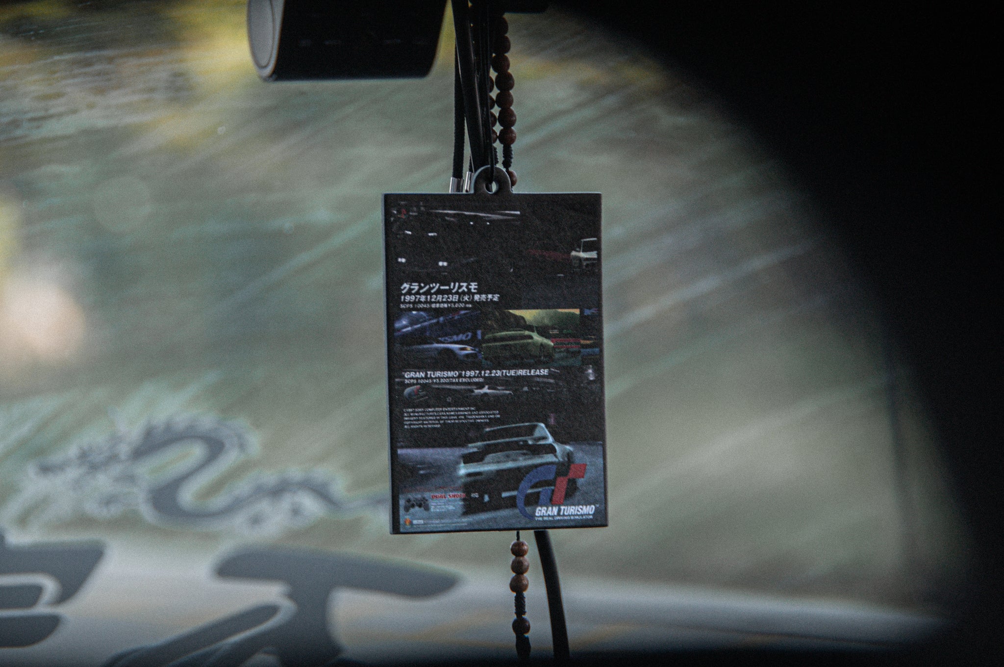 A close-up of a rearview mirror shows an Incompletegl Retro Gran Turismo! air freshener card with Japanese text and car images, hanging on a beaded string. Part of a dragon sticker is visible on the windshield.