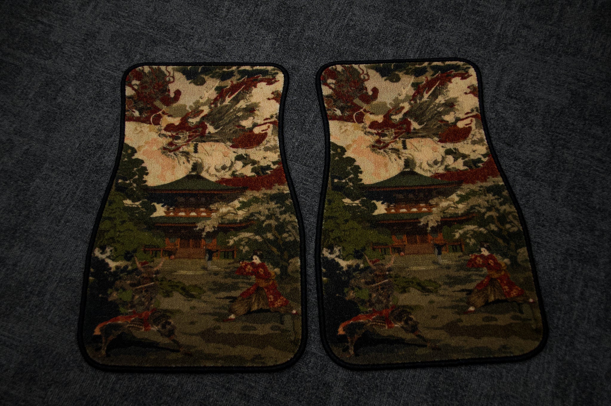 Two car floor mats from Incompletegl, called Ryujin x Raijin, 'The Palace Painting,' featuring intricate designs of a Japanese palace surrounded by trees and traditional buildings set against a gray background. The mats are placed side by side.