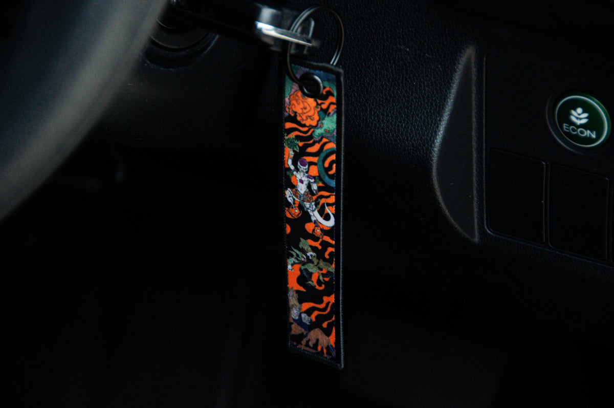 Close-up image of a car ignition with a key inserted, featuring a vibrant and stylish keychain from the Dragon Ball Z Super Pack! Bundle Pack by Incompletegl. The keychain displays an abstract mix of orange, black, green, and white designs that evoke iconic Dragon Ball Z aesthetics. The dashboard of the car is slightly visible with a button labeled "ECON" in green.