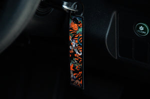 Close-up image of a car ignition with a key inserted, featuring a vibrant and stylish keychain from the Dragon Ball Z Super Pack! Bundle Pack by Incompletegl. The keychain displays an abstract mix of orange, black, green, and white designs that evoke iconic Dragon Ball Z aesthetics. The dashboard of the car is slightly visible with a button labeled "ECON" in green.