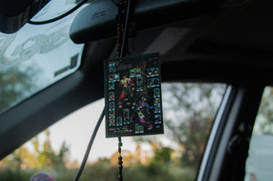 A close-up reveals an Incompletegl Retro Tekken!! air freshener hanging from the car's rearview mirror, with a background of blurred trees and a clear sky.