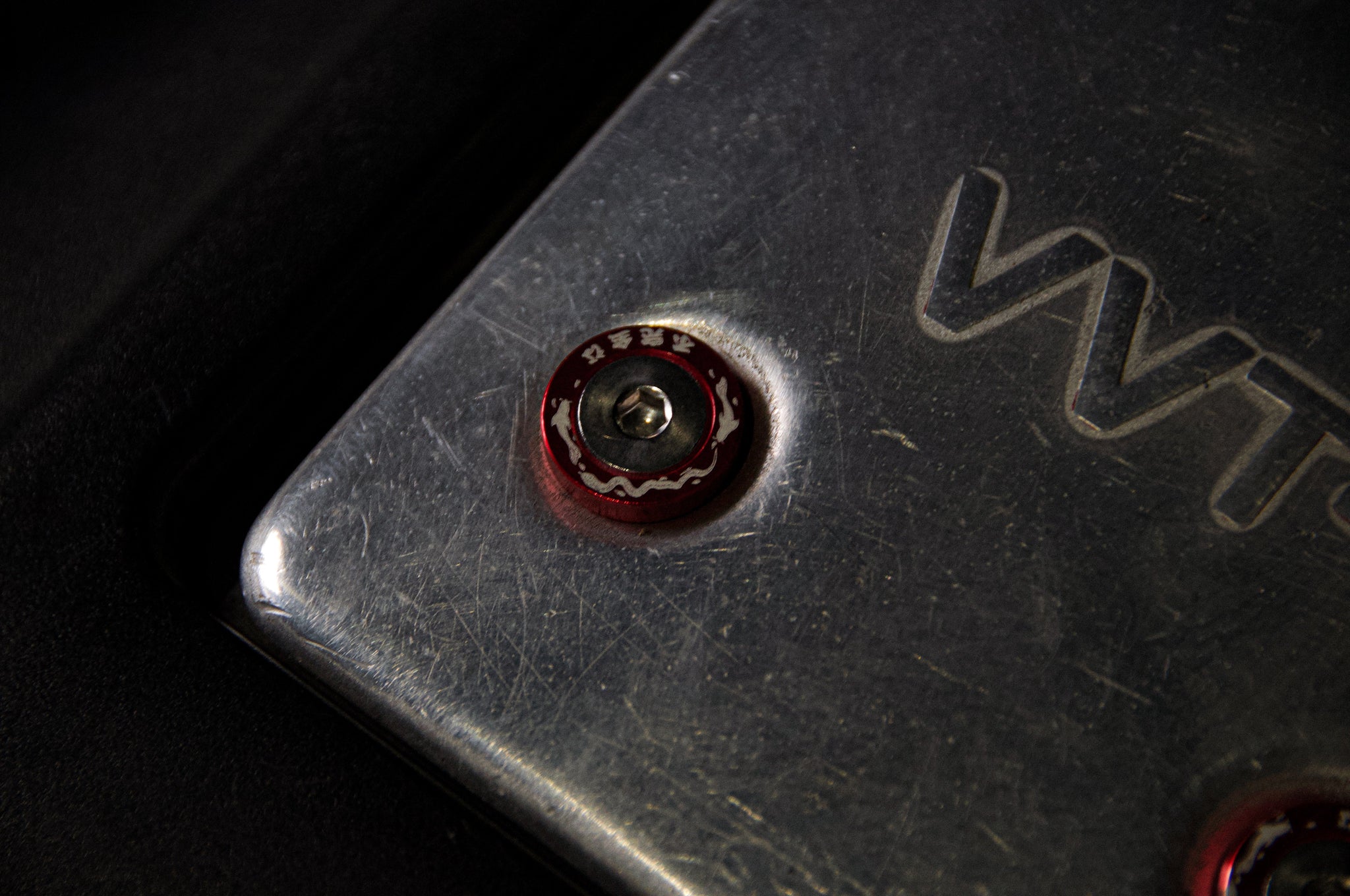 Close-up of a metal surface with the letters "VVTi" engraved on it. Near the center of the image, an Incompletegl Dress Up Bolt! | M6 Bolt is visible, characterized by its small, round shape, red hue, and white patterns accompanied by foreign characters. The photo highlights the texture and intricate details of the metal and this decorative bolt.