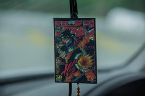 The Retro Naruto! air freshener by Incompletegl features an illustrated magazine cover with two animated characters in action poses, bold Japanese text, and vibrant colors against a blurred car dashboard and road background.