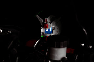 A close-up of the GENJI x RX78! | Limited Edition robot's animated face by Incompletegl in a dark setting, illuminated with white and red details, reminiscent of futuristic or science fiction headgear. Controls and dials, similar to those found in a JDM driving companion, are partially visible in the foreground.