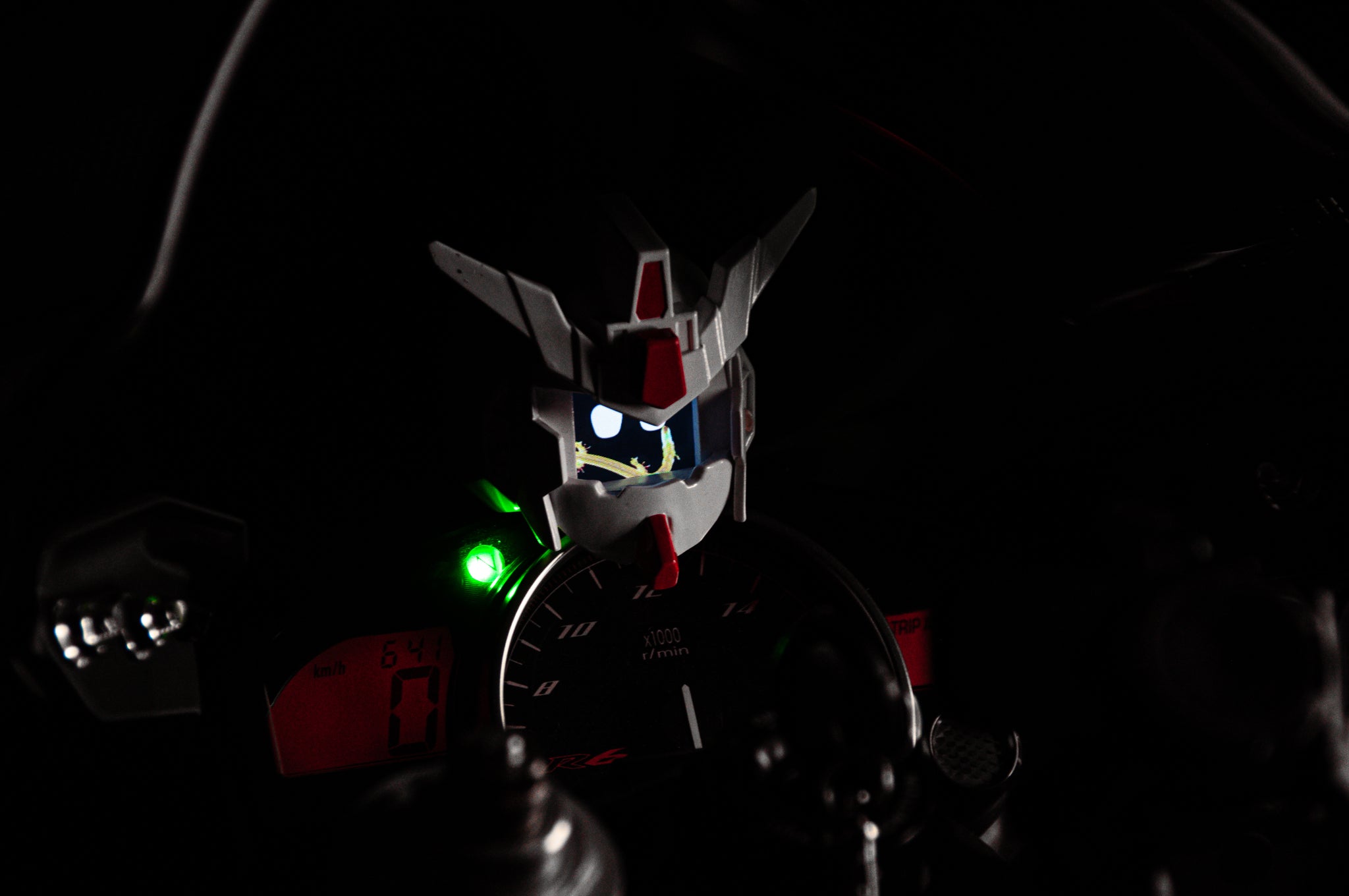 A close-up of a motorcycle dashboard with the GENJI x RX78! Limited Edition design by Incompletegl. The panel features an analog speedometer and digital displays, highlighted by an illuminated green light on the left side. The centerpiece is the iconic white and red Gundam head above the gauges—your ultimate JDM driving companion.