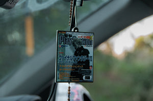 An Incompletegl Retro FFVII air freshener, inspired by vintage PlayStation magazine covers and Final Fantasy 7's magic, hangs from a car's rearview mirror with a blurred outdoor background seen through the windshield.