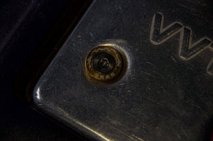 A close-up shot reveals a weathered metallic surface, centered around an M6 bolt from Dress Up Bolts! by Incompletegl. The single bolt displays slight rusting, while the surrounding area features noticeable scratches and wear. Partially legible text or engraving can be seen on the upper right side of the surface, all under low lighting conditions.