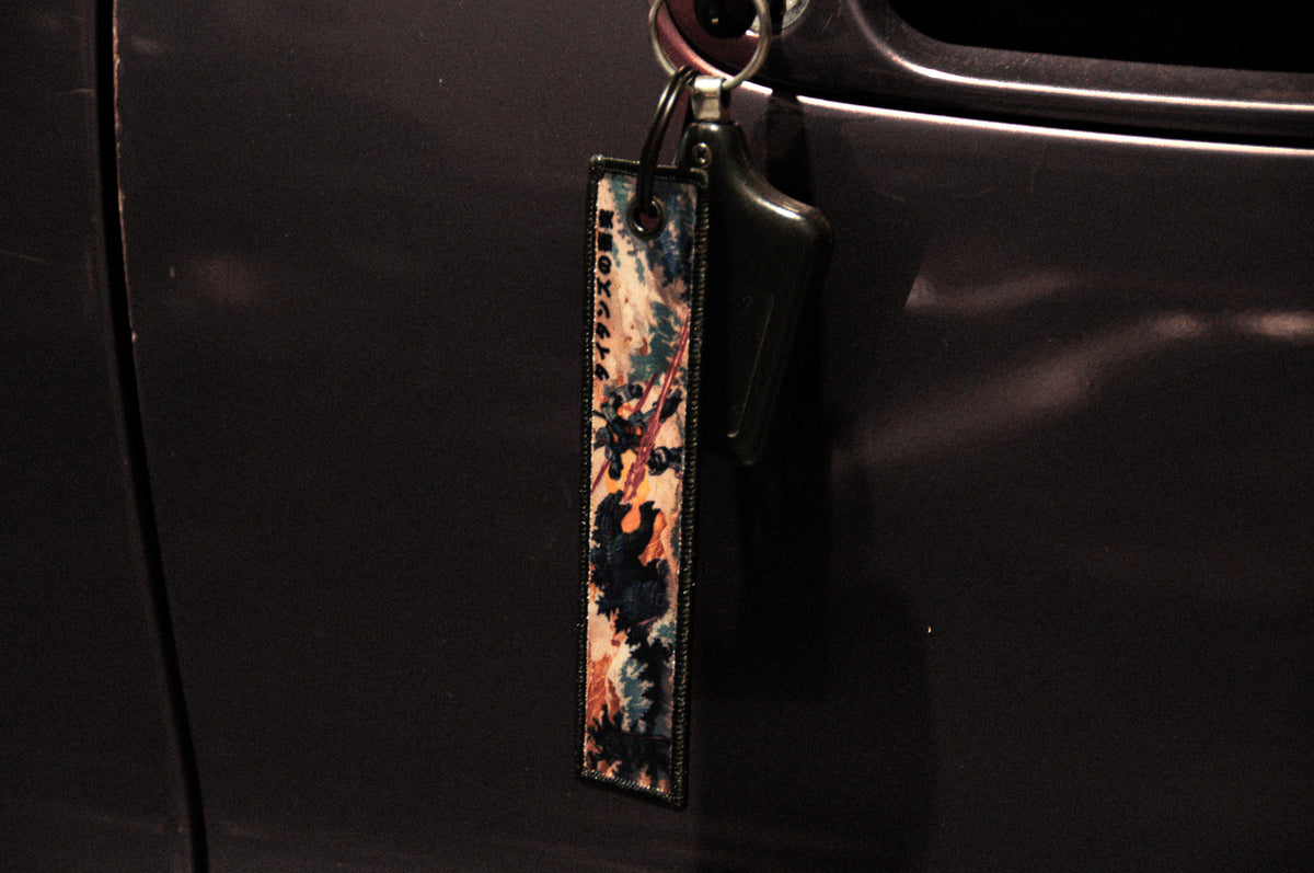 A close-up image of a black car door with a key inserted into the lock. Attached to the key is the Godzilla x Gundam Jet Tag by Incompletegl, showcasing a rectangular leather strap adorned with a colorful abstract design in shades of blue, red, and beige that captures Godzilla's cinematic flair. The image has a dark, moody tone.