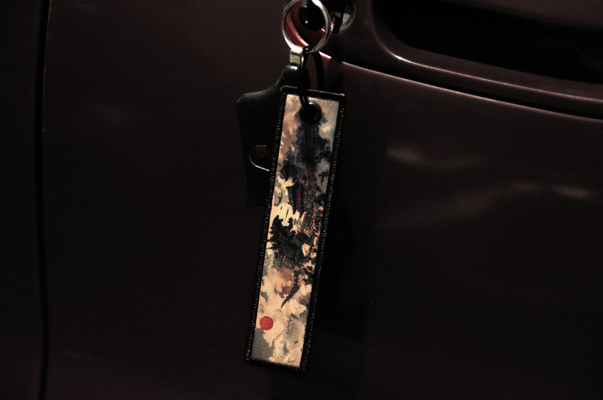 A close-up image of a car key inserted in the door lock of a dark-colored vehicle. Attached to the key is a rectangular keychain from Incompletegl, featuring a colorful abstract design inspired by Godzilla and Gundam aesthetics. The dimly lit background accentuates the key and the vibrant charm of the Jet Tag.