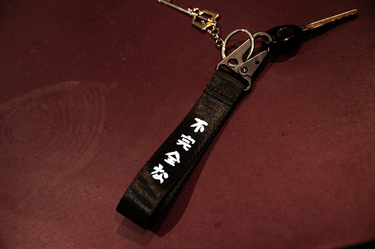 The Oni Mask Sword x Cherry Blossoms Carabiner Keychain by Incompletegl features a black canvas section enhanced with white Japanese characters, a metallic clip, and an added carabiner for convenience. This keychain, complete with a key, is displayed on a dark red surface, radiating an authentic Japanese aesthetic.