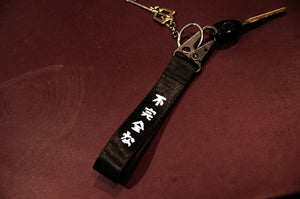 The Oni Mask Sword x Cherry Blossoms Carabiner Keychain by Incompletegl features a black canvas section enhanced with white Japanese characters, a metallic clip, and an added carabiner for convenience. This keychain, complete with a key, is displayed on a dark red surface, radiating an authentic Japanese aesthetic.