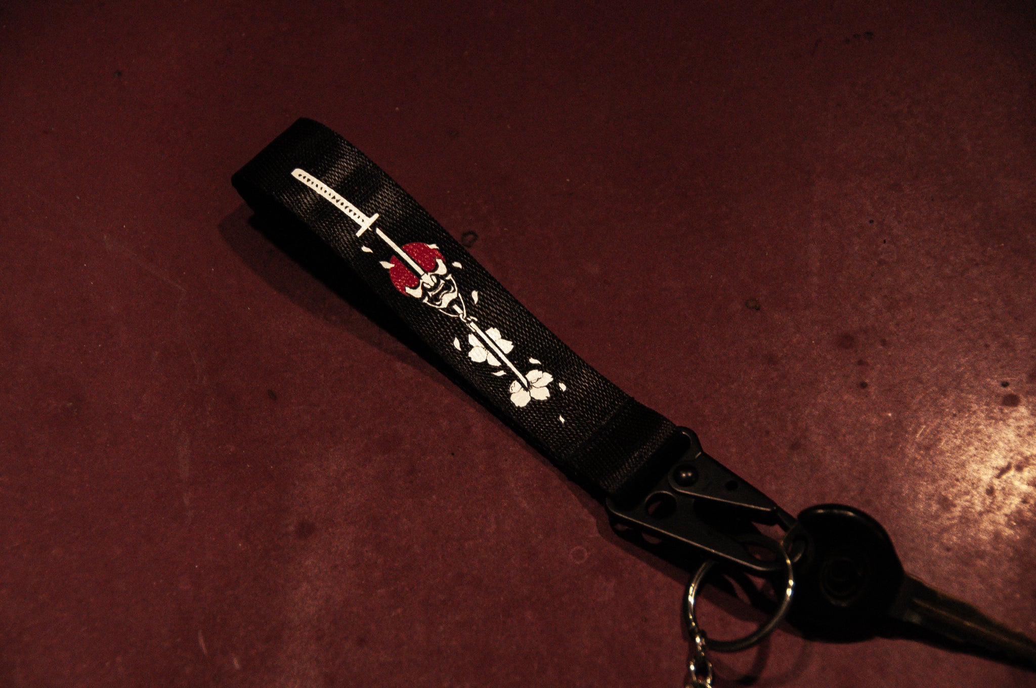 The Oni Mask Sword x Cherry Blossoms Carabiner Keychain by Incompletegl, featuring a black lanyard with a white design of a stylized skull and roses reminiscent of Japanese aesthetics, lies on a dark red surface. The lanyard has a metal clip and is attached to a key.