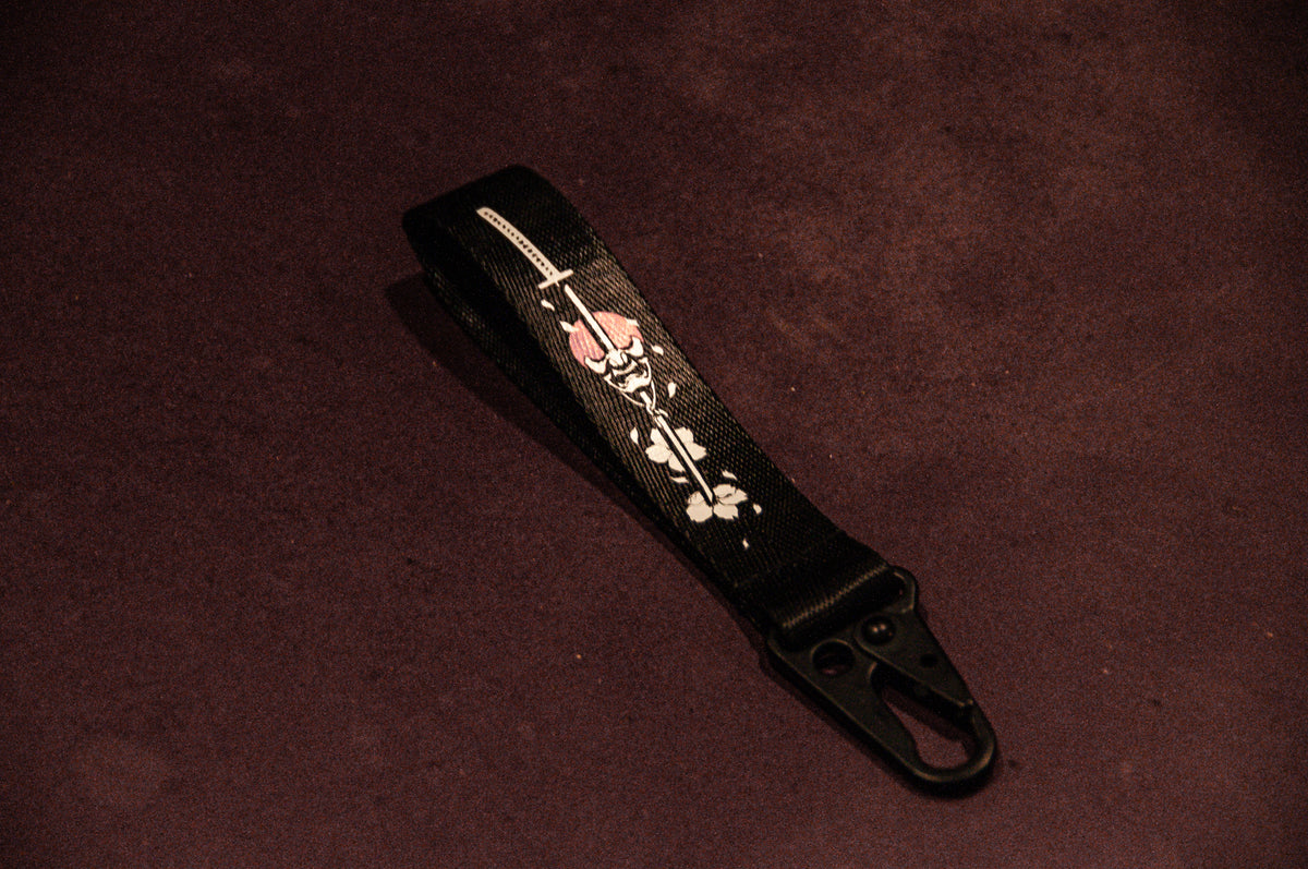 An Oni Mask Sword x Cherry Blossoms carabiner keychain from Incompletegl, featuring a metallic carabiner at one end and adorned with an intricate skull graphic wearing a red hat and crossbones, laid on a dark purple surface.