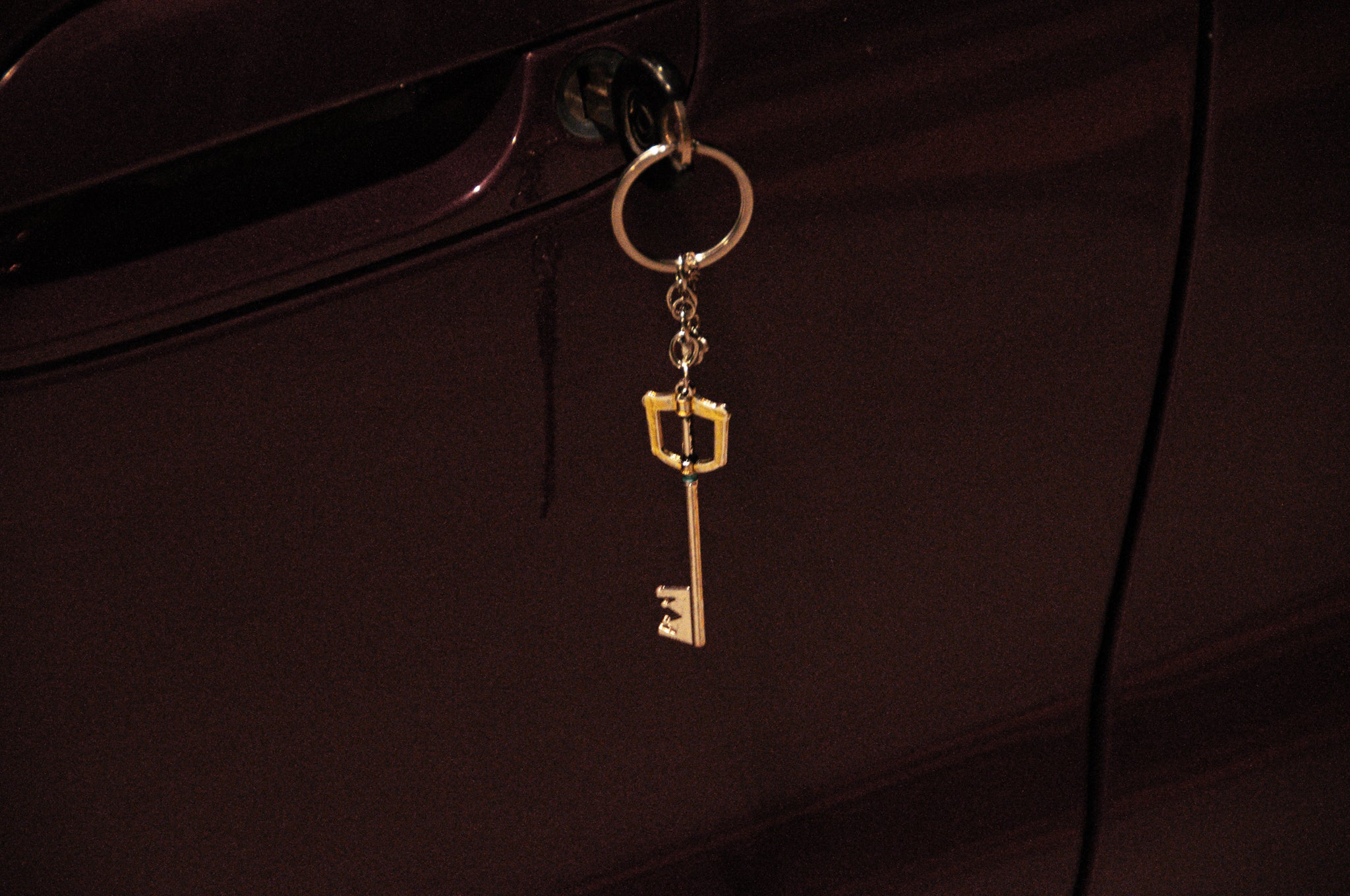 A Kingdom Hearts Key Blade keychain from Incompletegl dangles from the lock of a maroon-colored car door, featuring a decorative element that resembles a small, golden Keyblade reminiscent of designs crafted by Square Enix.