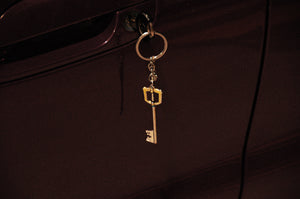 A Kingdom Hearts Key Blade keychain from Incompletegl dangles from the lock of a maroon-colored car door, featuring a decorative element that resembles a small, golden Keyblade reminiscent of designs crafted by Square Enix.