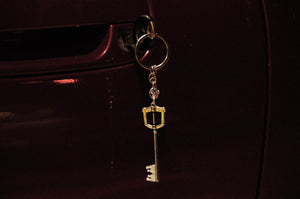 A Kingdom Hearts Key Blade keychain by Incompletegl dangles from the door lock of a maroon-colored car. The car's door handle is partially visible. The image has a dark, moody tone with minimal lighting, evoking the atmospheres characteristic of Kingdom Hearts.