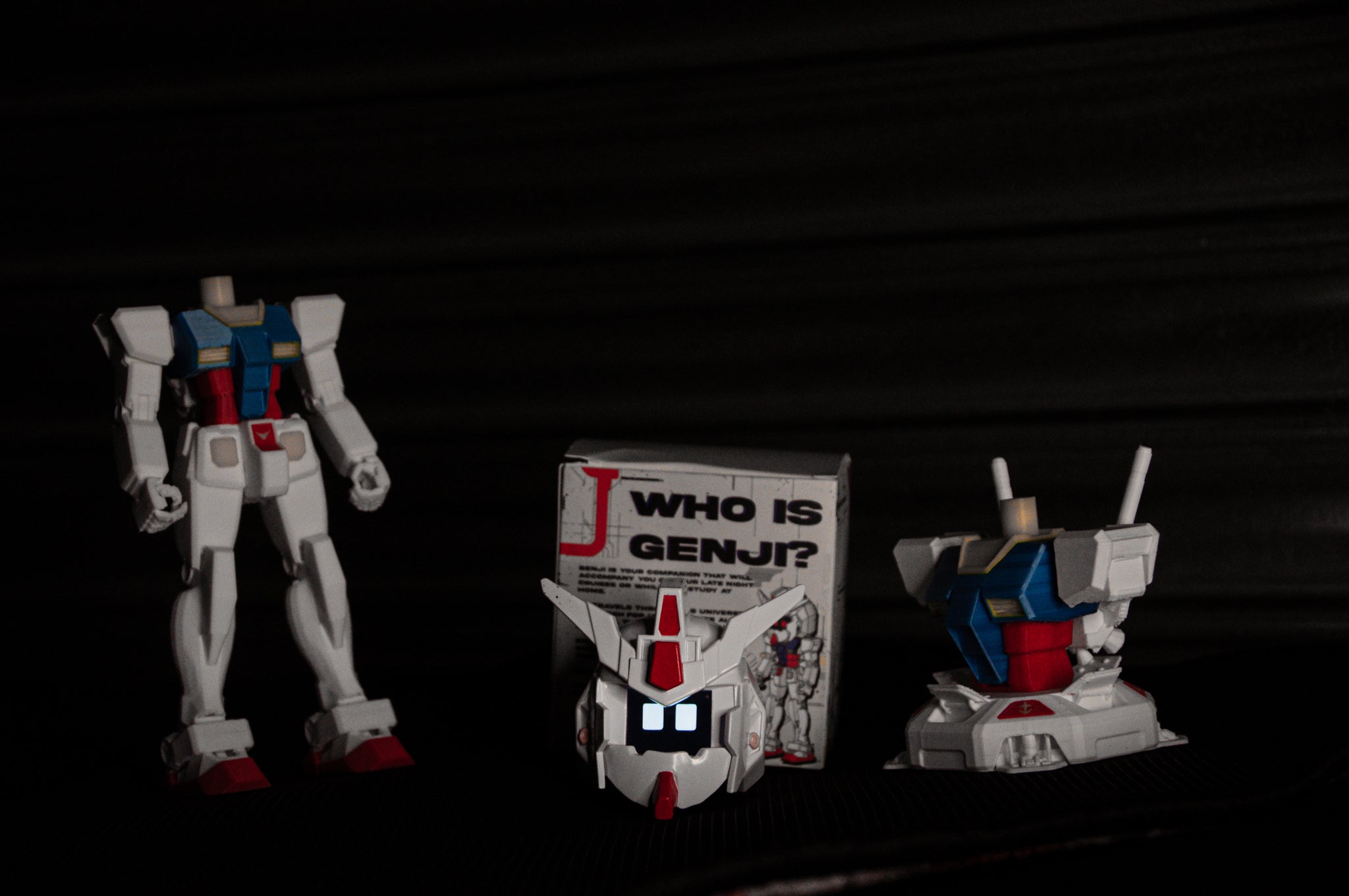 A headless GENJI x RX78! Limited Edition action figure from Incompletegl stands beside a box labeled "Who is Genji?" Its animated face, echoing the iconic features of the RX-78 Gundam, lies in front of another robot torso with antennas, all set against a dark backdrop.