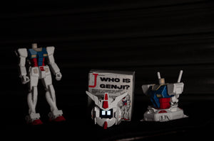 A headless GENJI x RX78! Limited Edition action figure from Incompletegl stands beside a box labeled "Who is Genji?" Its animated face, echoing the iconic features of the RX-78 Gundam, lies in front of another robot torso with antennas, all set against a dark backdrop.