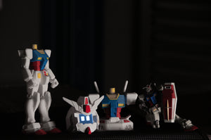 Four unassembled parts from the GENJI x RX78! Limited Edition model by Incompletegl are arranged on a dark surface. Each piece is a section of the iconic robot, showcasing blocky shapes and colors like white, blue, red, and yellow. Shadows enhance the details on these individual components.